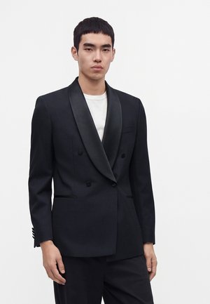 Jeune homme portant un blazer noir croisé avec des revers en satin sur une chemise blanche, debout devant un fond clair uni.