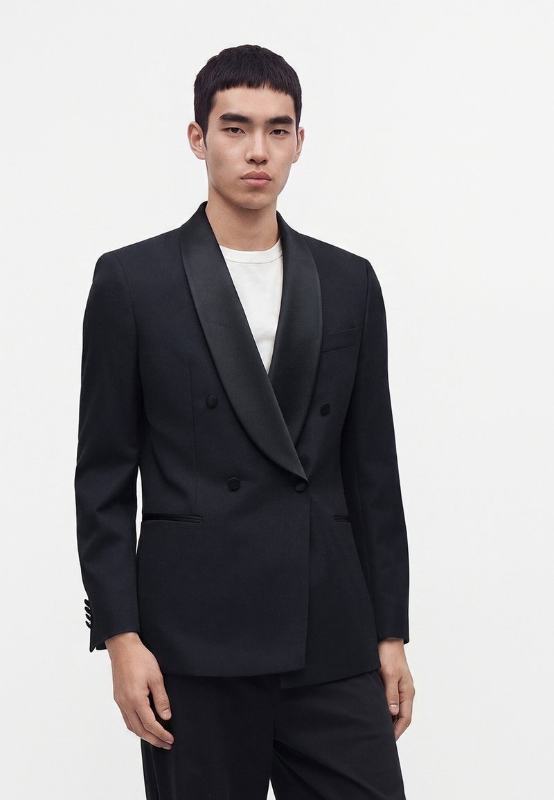 Jeune homme portant un blazer noir croisé avec des revers en satin sur une chemise blanche, debout devant un fond clair uni.
