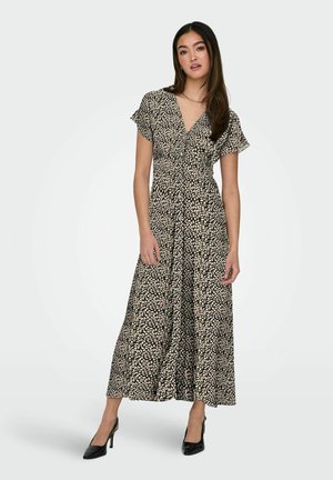 Kobieta stojąca, ubrana w midi sukienkę z krótkim rękawem w czarno-beżowy wzór liści oraz czarne buty na wysokim obcasie, na jednolitym tle.