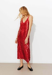 Robe longue rouge en sequins avec décolleté en V et fines bretelles, présentant une coupe ample et une texture scintillante. Assortie à des ballerines noires à pois.