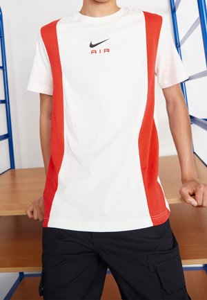 Osoba ubrana w biało-czerwony T-shirt Nike Air oraz czarne szorty typu cargo, opierająca się o drewniany stół z niebieską metalową ramą.