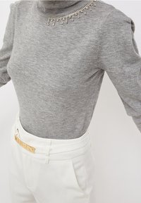 Grauer Rollkragenpullover mit dekorativen Strasssteinen am Halsausschnitt, kombiniert mit weißen, hochgeschnittenen Hosen und einer goldfarbenen Gürtelschnalle.