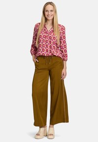 Blusa rosa con motivo a fantasia e maniche lunghe, abbinata a pantaloni larghi marroni con vita regolabile e ricami laterali. Scarpe beige con zeppa.