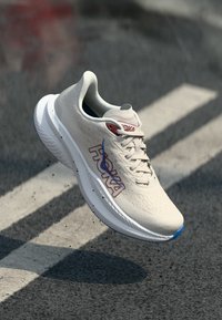 Sapatilha de corrida cinza claro com parte superior texturizada, detalhes em azul e vermelho, design de atacadores e uma sola branca grossa. Logótipo visível na lateral.