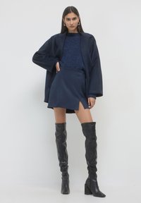 Donna che indossa un cappotto blu navy, un maglione a trama, una mini gonna con spacco frontale e stivali alti neri con tacco, in piedi su uno sfondo bianco.