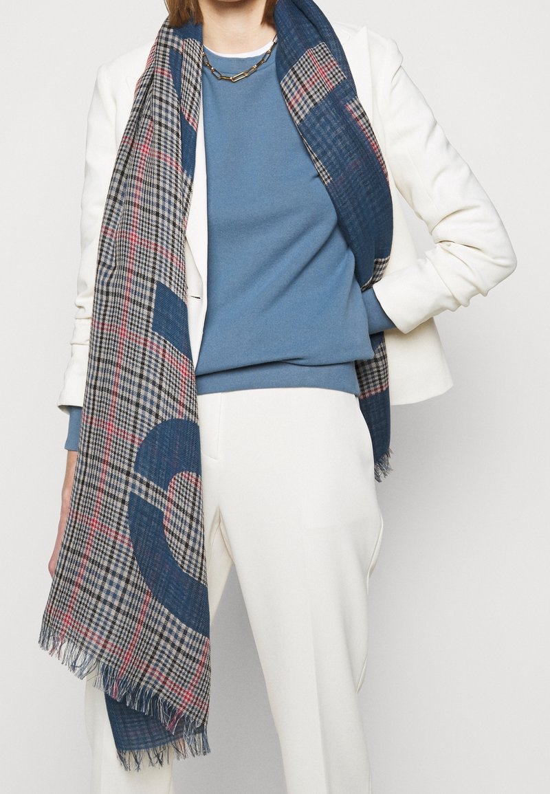 Personne portant un pantalon blanc, un blazer blanc, un pull bleu, un collier chaîne en or et une grande écharpe à carreaux avec des motifs bleus et rouges.