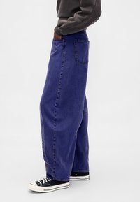 Ohlapne vijolične kavbojke iz jeansa s sproščenim krojem, s stranskimi žepi in zadnjim žepom, združene s črnimi platnenimi supergami.