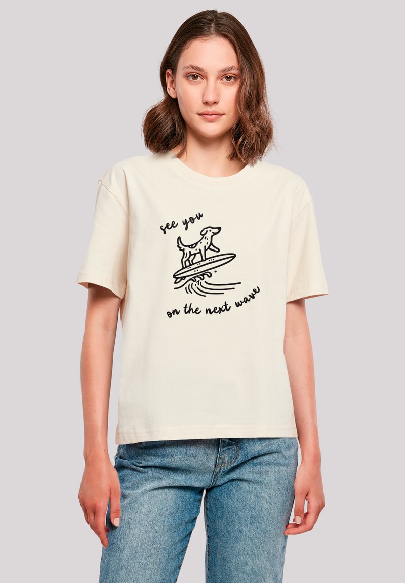 Beige Baumwoll-T-Shirt mit kurzen Ärmeln, das ein schwarzes Grafikdesign eines Hundes auf einem Surfbrett sowie den Text "Wir sehen uns auf der nächsten Welle" zeigt.