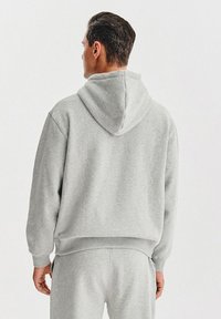 Szara, bezrękawnikowa bluza z kapturem, wykonana z miękkiego materiału, charakteryzująca się luźnym krojem i ściągaczami przy mankietach, widok z tyłu bez widocznych logo.