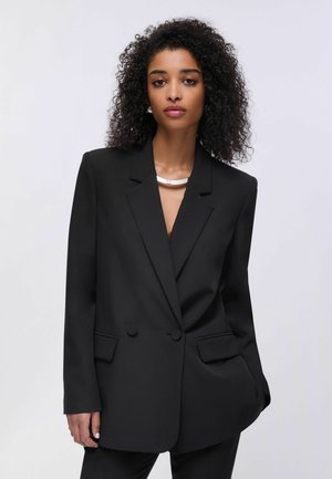 Kobieta o kręconych włosach, ubrana w czarną dwurzędową marynarkę i srebrny naszyjnik, stojąca na jednolitym jasnym tle.