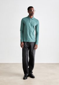 Cămașă polo cu mâneci lungi, de culoare teal, cu un mic logo, asortată cu pantaloni gri închis și pantofi negri, accesorizată cu ochelari de soare maro.