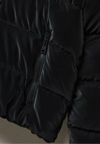 Černá prošívaná bunda v detailním záběru, ukazující vertikální zipovou kapsu na přední straně, zdůrazňující prošívání a lesklou texturu látky.