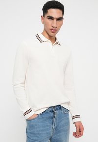 Polo de mangas compridas em tecido off-white com colarinho e punhos com riscas contrastantes, apresentando um discreto logo bordado no peito.