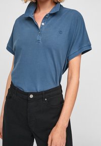 Polo s krátkým rukávem v modré barvě, vyrobený z texturované látky. Obsahuje knoflíkovou légu a malou značku. Vhodně doplněn o černé kalhoty s vysokým pasem.