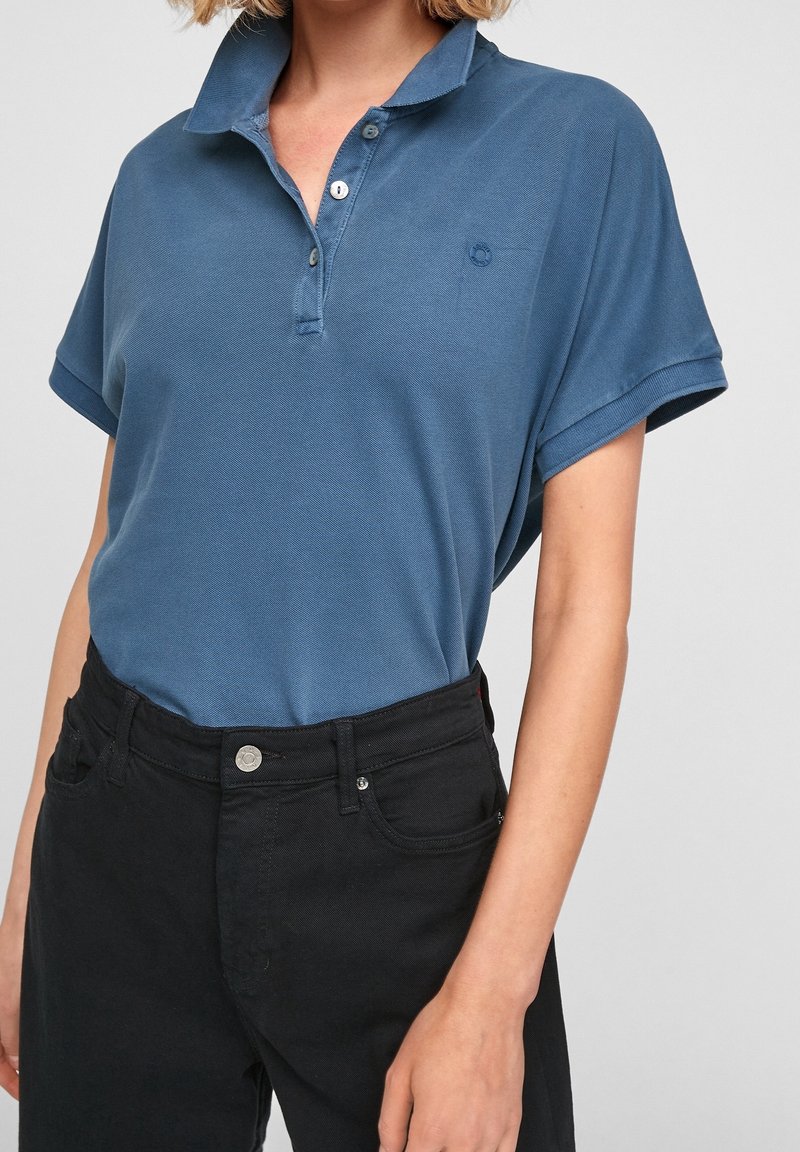 Polo s krátkým rukávem v modré barvě, vyrobený z texturované látky. Obsahuje knoflíkovou légu a malou značku. Vhodně doplněn o černé kalhoty s vysokým pasem.