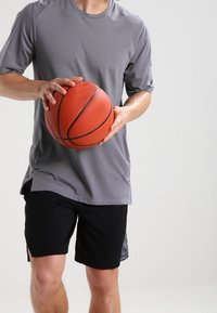 Camiseta atlética corta gris, pantalones cortos negros, sosteniendo un balón de baloncesto naranja con textura y costuras visibles, contra un fondo neutro.