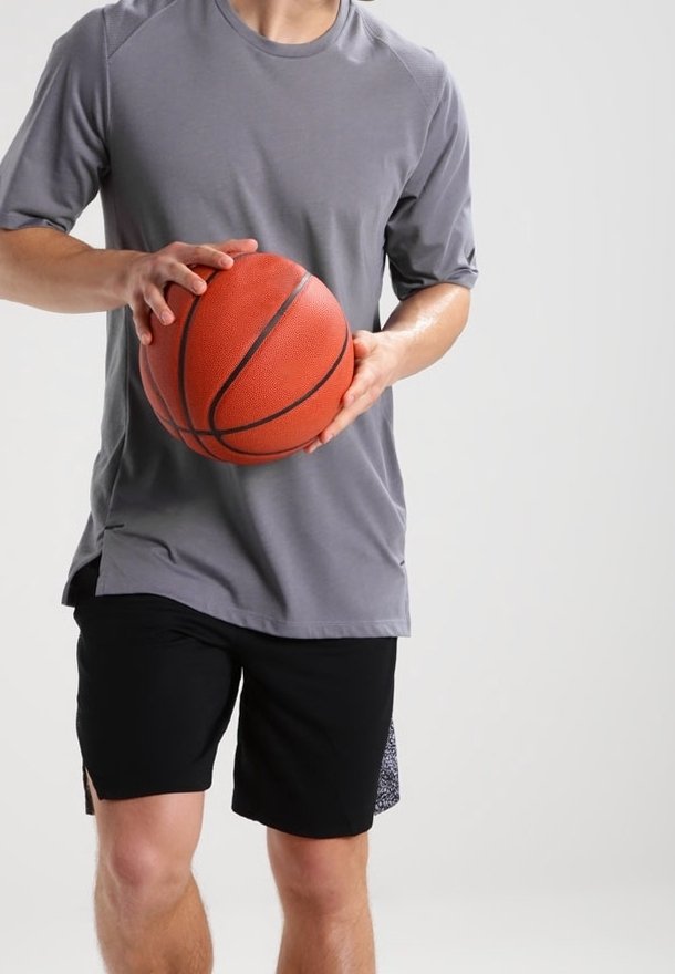 Camiseta atlética corta gris, pantalones cortos negros, sosteniendo un balón de baloncesto naranja con textura y costuras visibles, contra un fondo neutro.