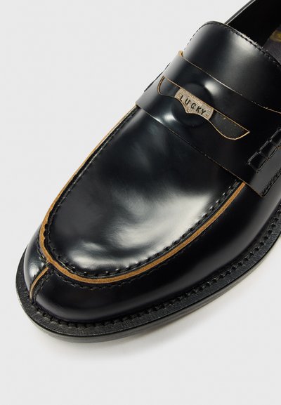Mocassin en cuir noir avec une sangle décorative portant le mot "LUCKY." Gros plan mettant en avant la brillance et les détails de fabrication.