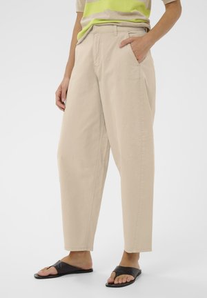 Personne portant un pantalon beige taille haute à jambes larges, une main dans la poche, associé à des sandales noires à enfiler et un haut partiellement rayé vert.