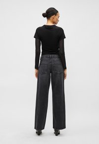 Žena s vlasmi v drdole, oblečená v čiernom vrstvenom dlhom rukáve, vysokým pásom a širokými tmavými džínsy a čiernymi lodičkami, otočená chrbtom na jednoliatom pozadí.