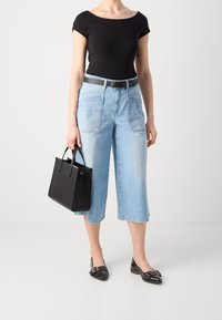 Žena v černém přiléhavém topu, světle modrých širokých cropped džínech, s černým opaskem a černými špičatými loafers, držící černou kabelku.