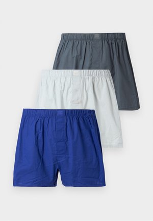 Lange, slanke man in witte shorts staat op blote voeten naast een donkergroene lederen fauteuil tegen een eenvoudige witte muur.