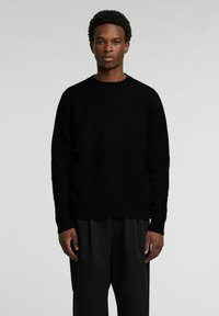 Schwarzer, gestrickter Pullover mit rundem Ausschnitt und langen Ärmeln, versehen mit einer strukturierten Oberfläche. Kombiniert mit lockeren schwarzen Hosen.