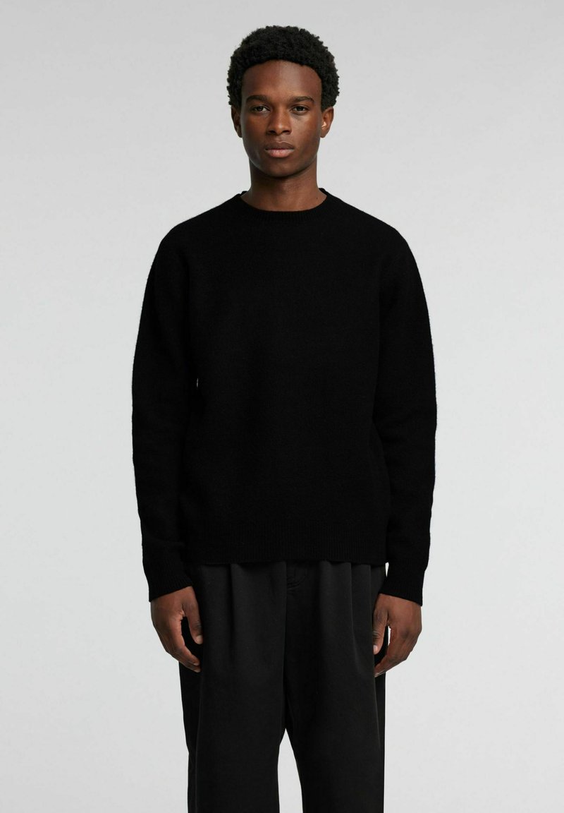 Schwarzer, gestrickter Pullover mit rundem Ausschnitt und langen Ärmeln, versehen mit einer strukturierten Oberfläche. Kombiniert mit lockeren schwarzen Hosen.