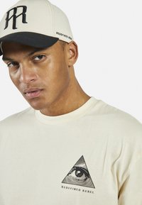 Cream-colored t-shirt with triangular black graphic featuring an eye and "REDEFINED REBEL" text; paired with a two-tone cap.