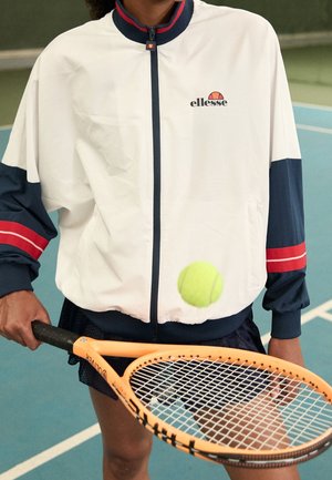 Persoon in een wit en marineblauw ellesse-jasje die een oranje tennisracket vasthoudt en een gele tennisbal slaat op een binnenbaan.