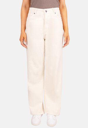 Frau trägt hoch taillierte, weit geschnittene, off-white Hose und weiße Turnschuhe, steht vor einem schlichten Hintergrund.