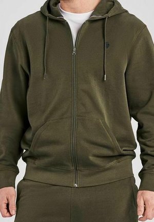 Mann trägt olivegrünen Kapuzenpullover mit Reißverschluss, Kordeln, Fronttaschen und passenden Hosen, teilweise sichtbar vom Hals bis zur Taille.