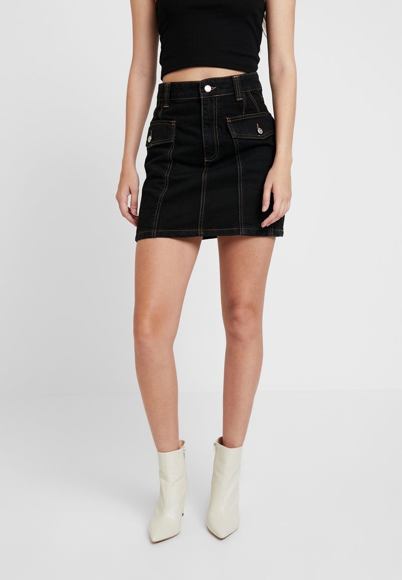Femme portant une mini-jupe noire taille haute avec des poches devant et des bottines blanches, debout devant un fond uni.