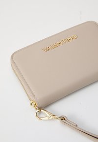 Beżowy skórzany portfel ze złotym zamkiem błyskawicznym i okuciami. Na froncie znajduje się złote logo "VALENTINO", do którego dołączony jest pasek na nadgarstek.