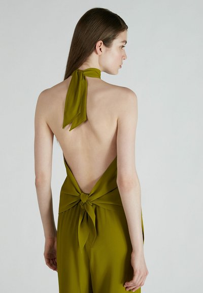 Femme aux longs cheveux bruns portant une combinaison dos nu olive verte avec des nœuds noués au cou et dans le bas du dos, de profil.
