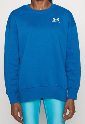 Henkilö, jolla on yllä sininen Under Armour -collegepaita ja vaaleansiniset urheiluleggingsit, seisoo yksivärisen taustan edessä.