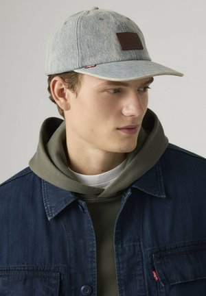 Young man wearing light gray denim cap, olive green hoodie, and dark blue denim jacket, looking to the side against a plain background.