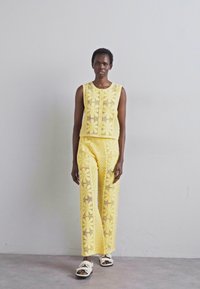 Ensemble en deux pièces au crochet jaune composé d'un haut sans manches avec des motifs floraux et d'un pantalon large, complété par des sandales blanches.