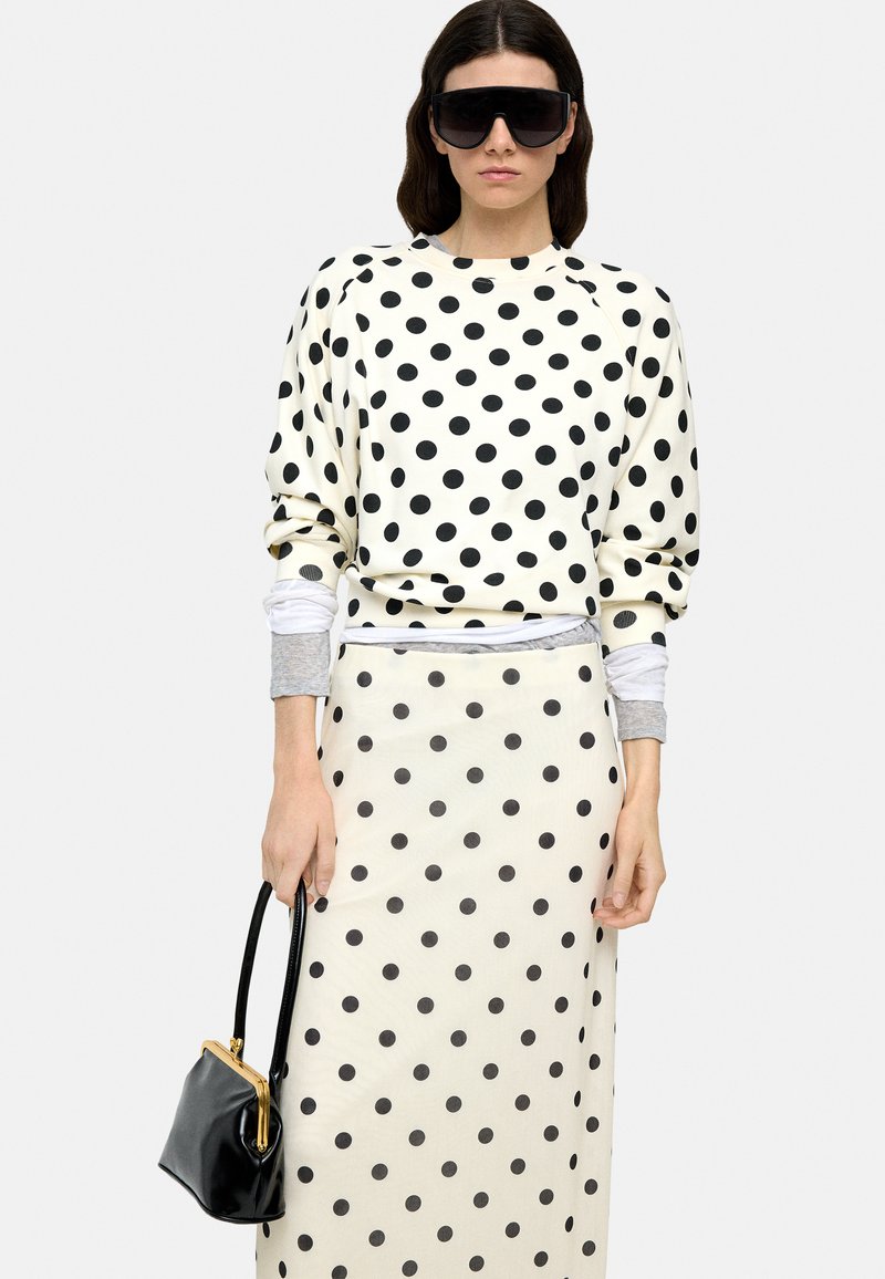 Biały sweatshirt w białe kropki i dopasowana spódnica, obie z elastycznego materiału. Modelka trzyma czarną skórzaną torebkę z złotym zapięciem. Czarne okulary przeciwsłoneczne.