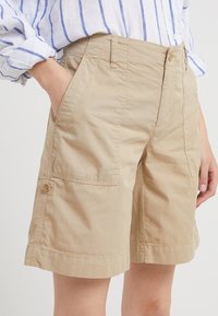 Pantaloni corti beige fino al ginocchio con ampie tasche frontali e un dettaglio con bottone laterale, indossati con una camicia a righe bianca e blu.