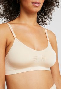 Bež wireless bralette izrađen od glatkog materijala. Ima nabran središnji deo, podesive trakice i meki elastični pojas za udobnost.