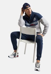 Dunkelblaue und graue Zip-Hoodie mit Logo, kombiniert mit dunkelblauen Trainingshosen. Das Model sitzt auf einem weißen Stuhl, trägt gemusterte Sneaker und eine dunkelblaue Cap.