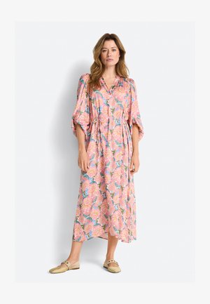 Kvinde iført en pink blomster-middellang kjole med puffede ærmer og beige slip-on sko stående mod en ensfarvet hvid baggrund.