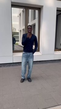 Person klädd i en marinblå cardigan, ljusblå jeans och svarta skor, stående på asfalt framför en modern byggnad med stora fönster.