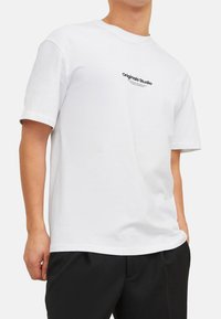 Weißes Baumwoll-T-Shirt mit kurzen Ärmeln, rundem Halsausschnitt und schwarzem "Originals Studio"-Text, der auf der Vorderseite gedruckt ist. Lässige Passform.