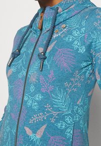 Sudadera azul con cremallera y patrones de flores y hojas en rosa y azul verdoso. Cuenta con capucha con cordón y cremallera metálica con detalles en madera.