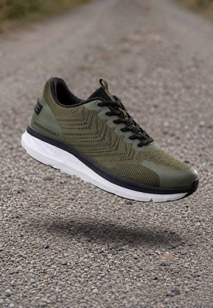 Scarpa da corsa verde oliva e nera con suola bianca etichettata Swedemount, che fluttua sopra un sentiero coperto di ciottoli.