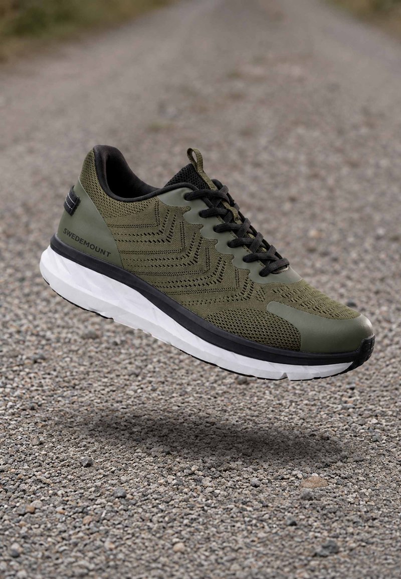 Chaussure de course vert olive et noir avec semelle blanche, portant l’étiquette Swedemount, flottant au-dessus d’un chemin recouvert de galets.