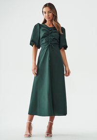 Dark green, satin maxi dress with puff sleeves, ruched bodice, tie detail, and a flowing skirt, paired with strappy heels.