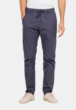 Hombre con pantalones azul marino con cordón y zapatillas beige, de pie con las manos parcialmente en los bolsillos, frente a un fondo sencillo.